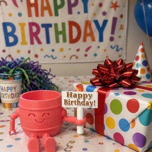Puede incluir: Un recipiente en forma de barril de color coral con brazos y piernas, que sostiene un letrero que dice "Happy Birthday!". Un regalo envuelto con un lazo rojo y un patr&oacute;n de lunares de colores est&aacute; cerca. Una pancarta con "Happy Birthday!" est&aacute; en el fondo.
