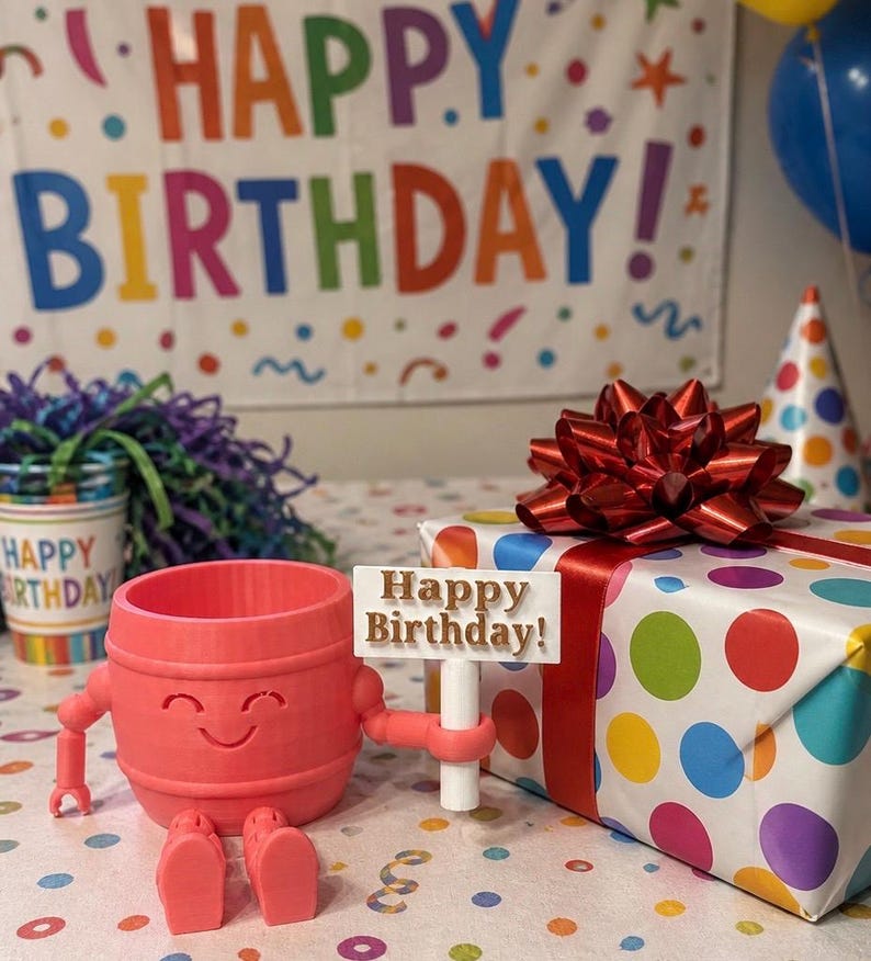 Puede incluir: Un recipiente en forma de barril de color coral con brazos y piernas, que sostiene un letrero que dice "Happy Birthday!". Un regalo envuelto con un lazo rojo y un patr&oacute;n de lunares de colores est&aacute; cerca. Una pancarta con "Happy Birthday!" est&aacute; en el fondo.