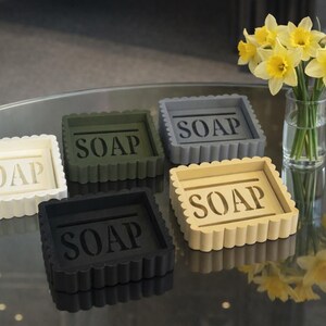 May include: Five square soap dishes in cream, olive green, gray, black, and tan, each with the word "SOAP" in the center. The dishes have a scalloped edge. A vase of yellow daffodils sits nearby.