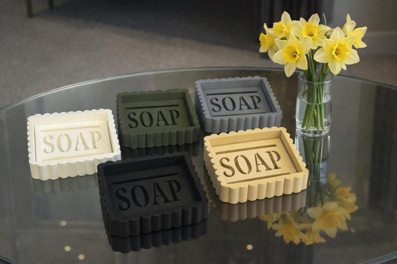 May include: Five square soap dishes in cream, olive green, gray, black, and tan, each with the word "SOAP" in the center. The dishes have a scalloped edge. A vase of yellow daffodils sits nearby.