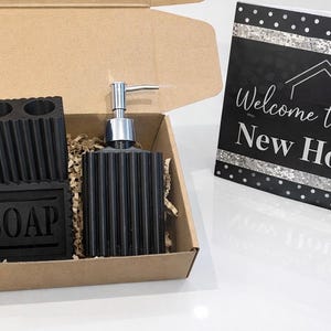May include: A black bathroom set in a cardboard box, including a soap dispenser, toothbrush holder, and soap dish. The soap dish is labeled "SOAP". A card with the text "Welcome to your New Home" is also included.