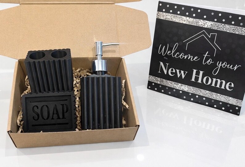 May include: A black bathroom set in a cardboard box, including a soap dispenser, toothbrush holder, and soap dish. The soap dish is labeled "SOAP". A card with the text "Welcome to your New Home" is also included.