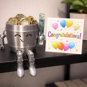 May include: A silver-colored, smiling barrel-shaped figurine filled with gold coins and a rolled-up bill. The figurine has robotic arms and legs, with silver boots. A greeting card with colorful balloons and the word "Congratulations!" is next to the figurine.