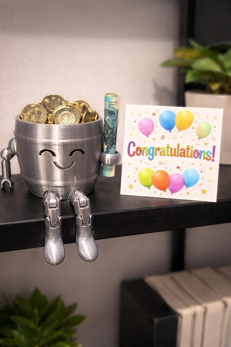 May include: A silver-colored, smiling barrel-shaped figurine filled with gold coins and a rolled-up bill. The figurine has robotic arms and legs, with silver boots. A greeting card with colorful balloons and the word "Congratulations!" is next to the figurine.