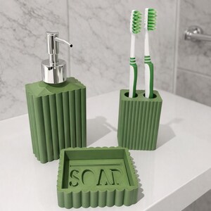 May include: A green bathroom set includes a soap dispenser with a silver pump, a toothbrush holder with two toothbrushes, and a soap dish with the word "SOAP". The set has a ribbed design.