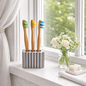 May include: Three bamboo toothbrushes with green, yellow, and blue bristles are in a silver holder. The holder sits on a windowsill next to a vase of white flowers and a white towel. The toothbrushes are a natural, eco-friendly option.