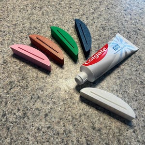 May include: A collection of colorful toothpaste tube squeezers in pink, brown, green, and blue, alongside a tube of Colgate toothpaste. A white tube squeezer is also present. The squeezers are designed to help get the most out of toothpaste tubes.
