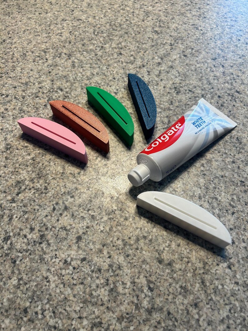 May include: A collection of colorful toothpaste tube squeezers in pink, brown, green, and blue, alongside a tube of Colgate toothpaste. A white tube squeezer is also present. The squeezers are designed to help get the most out of toothpaste tubes.