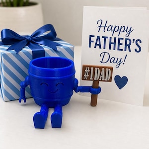 Puede incluir: Una maceta azul en forma de barril con brazos y piernas, sosteniendo un letrero que dice "#1DAD". Una tarjeta blanca con "Happy FATHER'S Day!" en escritura azul est&aacute; al lado, junto con una caja de regalo a rayas azules y blancas con un lazo azul.