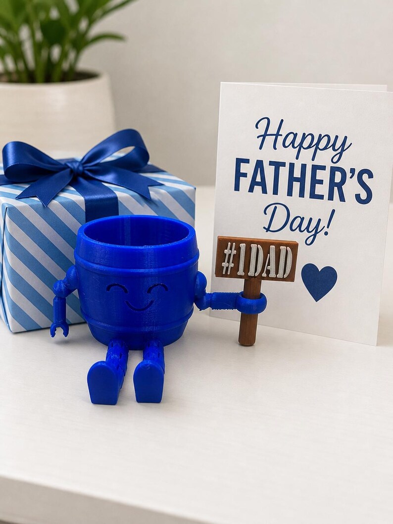 Puede incluir: Una maceta azul en forma de barril con brazos y piernas, sosteniendo un letrero que dice "#1DAD". Una tarjeta blanca con "Happy FATHER'S Day!" en escritura azul est&aacute; al lado, junto con una caja de regalo a rayas azules y blancas con un lazo azul.