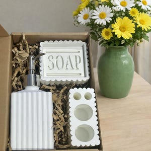 May include: A white bathroom set includes a soap dispenser, soap dish with "SOAP" text, and toothbrush holder. The set is in a cardboard box with paper filler. A green vase holds yellow and white daisies.