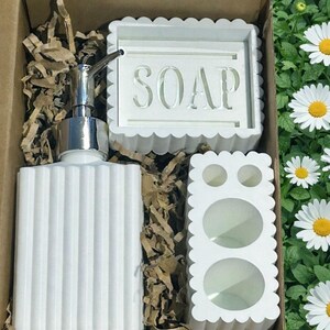 May include: A white bathroom set in a cardboard box, including a ribbed soap dispenser with a silver pump, a soap dish with the word "SOAP", and a toothbrush holder. The set has a scalloped edge design.