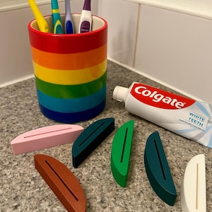 May include: A rainbow-striped toothbrush holder with four toothbrushes and a tube of Colgate toothpaste. Several colorful, wooden toothbrush stands in shades of pink, brown, green, and teal are also present.