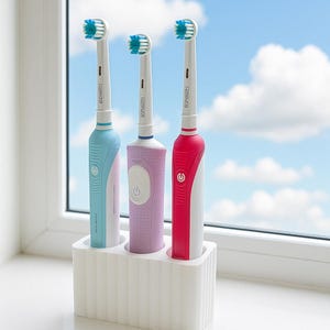 Puede incluir: Tres cepillos de dientes el&eacute;ctricos en un soporte blanco. Los cepillos de dientes son de color azul claro, lavanda y rojo, con cabezales blancos y azules. El soporte est&aacute; sobre un alf&eacute;izar blanco con cielo azul y nubes al fondo.