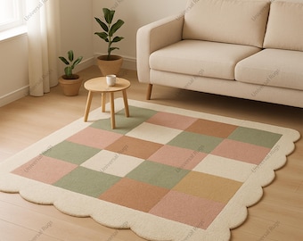 May include: A rectangular area rug with a scalloped edge. The rug features a checkerboard pattern in shades of pink, green, beige, and brown. The border is a solid cream color. The rug is placed on a light-colored wooden floor.
