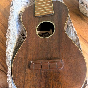 May include: A vintage ukulele with a dark brown wooden body and a round sound hole. The instrument has a light-colored fretboard and bridge. The ukulele is resting in a decorative, off-white, textured bowl.