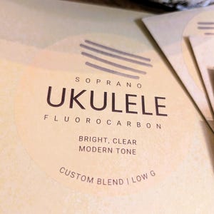May include: Close-up of a light tan ukulele string package. The text on the package reads "Soprano Ukulele Fluorocarbon" with additional text: "Bright, Clear, Modern Tone" and "Custom Blend | Low G".