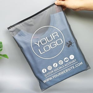 May include: A blue crew neck t-shirt inside a clear plastic bag with a black zipper. The bag has a white circular logo that says "YOUR LOGO" and social media icons. The website address is also printed on the bag.