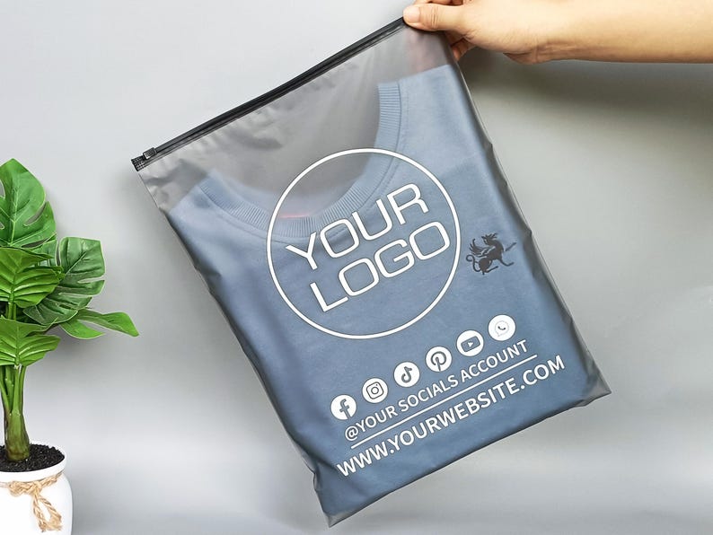 May include: A blue crew neck t-shirt inside a clear plastic bag with a black zipper. The bag has a white circular logo that says "YOUR LOGO" and social media icons. The website address is also printed on the bag.