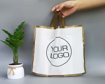 Op de afbeelding: Witte boodschappentas met gouden rand en handvatten. De tas heeft een zwart cirkelvormig logo met de tekst "YOUR LOGO". Links staat een kleine potplant met groene bladeren. De tas wordt door een hand vastgehouden.