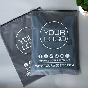 May include: Two gray plastic packaging bags with black zippers. One bag contains a dark blue shirt. The bags have a white circular logo with the text "YOUR LOGO". Below the logo are social media icons and a website address.