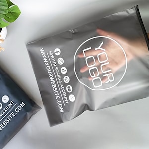 May include: Two gray poly mailer bags with a white logo and social media icons. One bag is held open by a hand. The text on the bags includes "YOUR LOGO", "@YOUR SOCIALS ACCOUNT", and "WWW.YOURWEBSITE.COM".