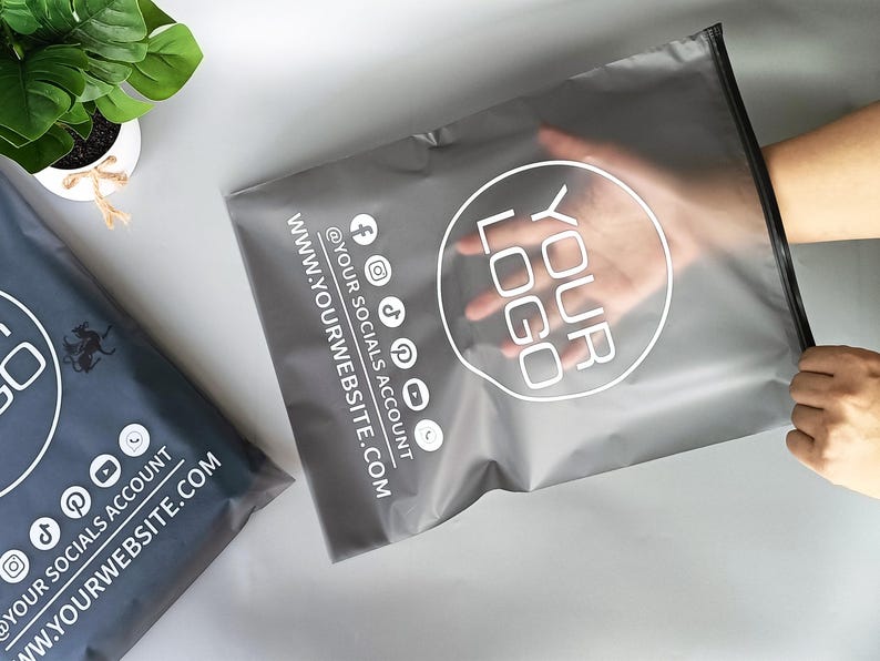 May include: Two gray poly mailer bags with a white logo and social media icons. One bag is held open by a hand. The text on the bags includes "YOUR LOGO", "@YOUR SOCIALS ACCOUNT", and "WWW.YOURWEBSITE.COM".