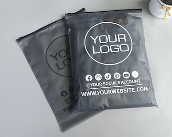 May include: Two gray poly mailer bags with black zippers. Each bag features a white circular logo with the text "YOUR LOGO". Below the logo are social media icons and the text "@YOUR SOCIALS ACCOUNT WWW.YOURWEBSITE.COM".