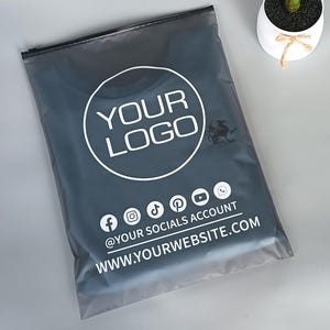 May include: A dark gray poly mailer bag with a white circular logo that says "YOUR LOGO". Below the logo are social media icons and the text "@YOUR SOCIALS ACCOUNT WWW.YOURWEBSITE.COM". A small plant in a white pot is in the background.
