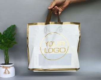 Op de afbeelding: Witte boodschappentas met gouden rand en handvatten, vastgehouden door een hand. De tas heeft een gouden cirkel met de woorden "YOUR LOGO" in goud gedrukt. De tas staat op een grijze achtergrond.