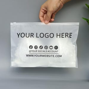 May include: A clear plastic bag with a zip closure, held by a hand. The bag displays the text "YOUR LOGO HERE", social media icons, and a website address in black. The background is a solid grey.