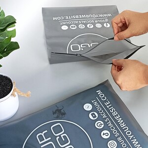 May include: Two gray packaging envelopes with a zipper closure are shown. The envelopes have a white logo and social media icons. Text on the envelope reads "@YOUR SOCIALS ACCOUNT" and "WWW.YOURWEBSITE.COM". A potted plant is in the background.