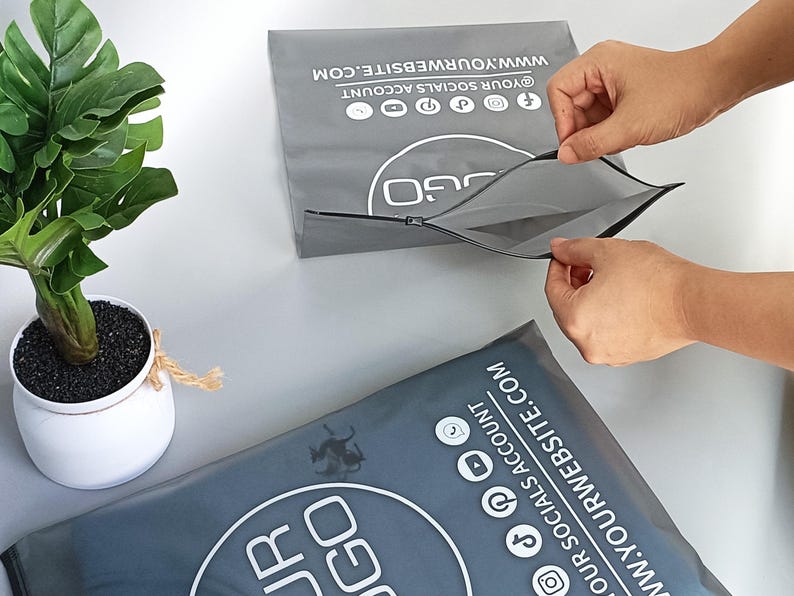 May include: Two gray packaging envelopes with a zipper closure are shown. The envelopes have a white logo and social media icons. Text on the envelope reads "@YOUR SOCIALS ACCOUNT" and "WWW.YOURWEBSITE.COM". A potted plant is in the background.