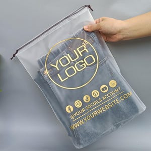 Può includere: Borsa trasparente in plastica con coulisse contenente jeans blu piegati. La borsa presenta un logo circolare dorato con la scritta "YOUR LOGO" e icone di social media. Il testo "@YOUR SOCIALS ACCOUNT" e "WWW.YOURWEBSITE.COM" sono anch'essi stampati in oro.