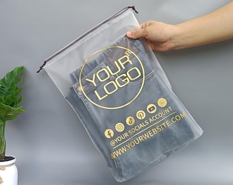 May include: Clear plastic drawstring bag containing folded blue jeans. The bag features a gold circular logo with the text "YOUR LOGO" and social media icons. The text "@YOUR SOCIALS ACCOUNT" and "WWW.YOURWEBSITE.COM" are also printed in gold.