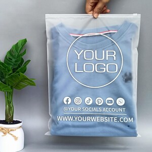 May include: A light blue crew neck sweatshirt in a clear plastic bag. The bag features a white circle with the text "YOUR LOGO" and social media icons. Below the logo is the text "@YOUR SOCIALS ACCOUNT" and "WWW.YOURWEBSITE.COM".