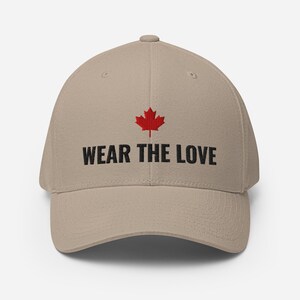 May include: A beige baseball cap with a curved peak. The cap displays a red maple leaf emblem above the black text "WEAR THE LOVE". The hat is a neutral colour and suitable for various outfits.