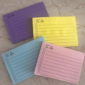 May include: Four rectangular notepads in purple, yellow, blue, and pink. Each notepad has horizontal lines and a column of circles on the left side. The words "To Do" are printed in black script at the top of each notepad.