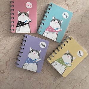 May include: Four spiral-bound notebooks in pink, blue, purple, and yellow. Each notebook features a cartoon cat with a fish and the word "MEW" in a speech bubble. The cat wears a scarf in a different colour on each notebook.