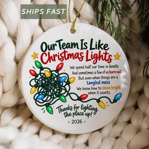 May include: A round white ceramic ornament featuring a graphic of tangled multi-coloured Christmas lights and the text: Our Team Is Like Christmas Lights, We spend half our time in knots, And sometimes a few of us burn out, But even when things are a tangled mess, We know how to shine bright when it counts, Thanks for lighting the place up!, - 2026 -.