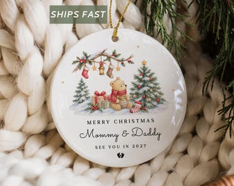 May include: A round white ceramic Christmas ornament featuring a watercolour illustration of a bear with a honey pot, a Christmas tree, and gifts. The text reads MERRY CHRISTMAS, Mommy & Daddy, SEE YOU IN 2027, with a small baby footprint icon.