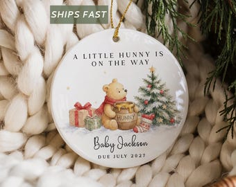 May include: A round white ceramic ornament featuring a bear holding a honey pot, a Christmas tree, and gifts. Text reads: A LITTLE HUNNY IS ON THE WAY, Baby Jackson, DUE JULY 2027. The ornament hangs from a gold string against a white knit background.