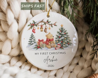 May include: A round white ceramic ornament featuring a festive illustration of a bear with a honey pot, a Christmas tree, and gifts. The text reads MY FIRST CHRISTMAS, the name Archie, and the year 2026. It hangs from a gold string.