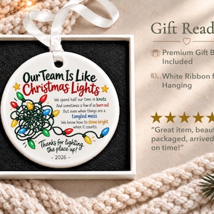 May include: A round white ceramic ornament with a white ribbon, featuring a graphic of tangled multi-coloured Christmas lights and the text: Our Team Is Like Christmas Lights, We spend half our time in knots, And sometimes a few of us burn out, But even when things are a tangled mess, We know how to shine bright when it counts, Thanks for lighting the place up, - 2026 -.