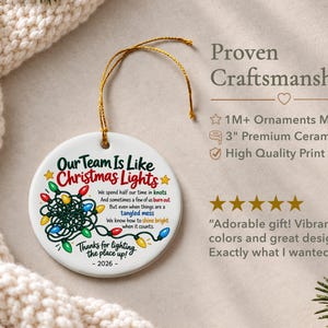May include: A round white ceramic ornament with a gold hanging string. The design features a graphic of tangled multi-coloured Christmas lights and text that reads: Our Team Is Like Christmas Lights, 2026, and other phrases. It measures 7.62 cm.