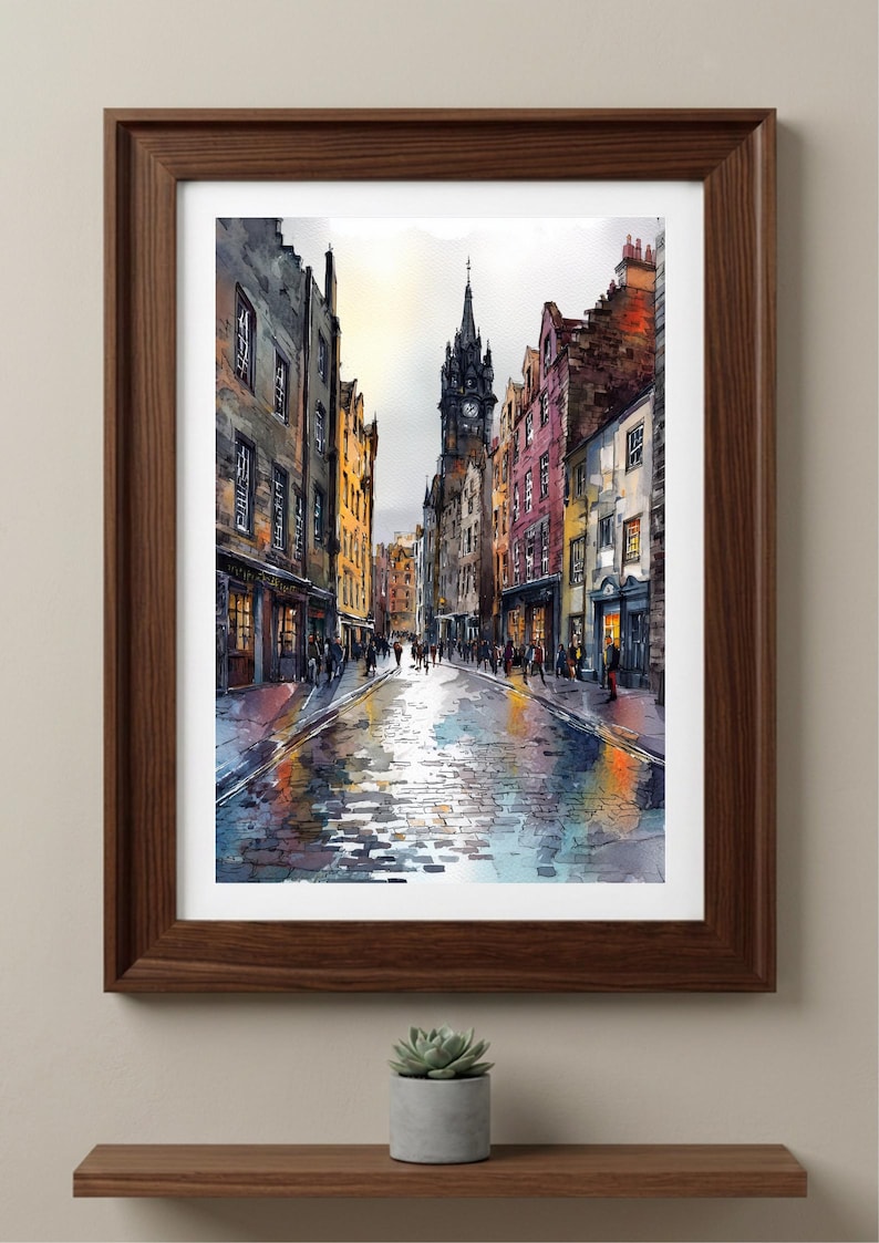 May include: A framed watercolor print depicting a narrow European cobblestone street lined with multi-story buildings in shades of yellow, red, and grey. A tall clock tower stands in the distance under a bright sky, with reflections on the wet ground.