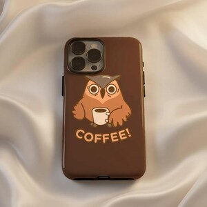 Può includere: Custodia per telefono marrone con un design a gufo dei cartoni animati che tiene una tazza di caffè. Il gufo è marrone e beige con grandi occhi. La parola "COFFEE!" è stampata in marrone sotto. Su sfondo di raso bianco.