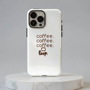Puede incluir: Funda de teléfono blanca con módulo de cámara negro y detalles negros. La funda presenta las palabras "coffee. coffee. coffee. loop." en marrón, fuente pixelada. La funda del teléfono se muestra en un soporte de mármol blanco.