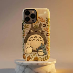 Könnte beinhalten: Eine Handyhülle mit einer Cartoon-Illustration von Totoro und anderen Charakteren. Die Hülle hat einen hellen Hintergrund mit einem Design aus Blättern und Eicheln. Die Charaktere befinden sich in der Mitte der Hülle, mit einem großen grauen Totoro und zwei kleineren Kreaturen.