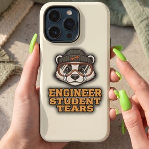 May include: A cream phone case with a cartoon badger design. The badger wears a beanie and goggles, with tears. The text "ENGINEER STUDENT TEARS" is in orange and yellow. The phone case is held by a person with long, green nails.